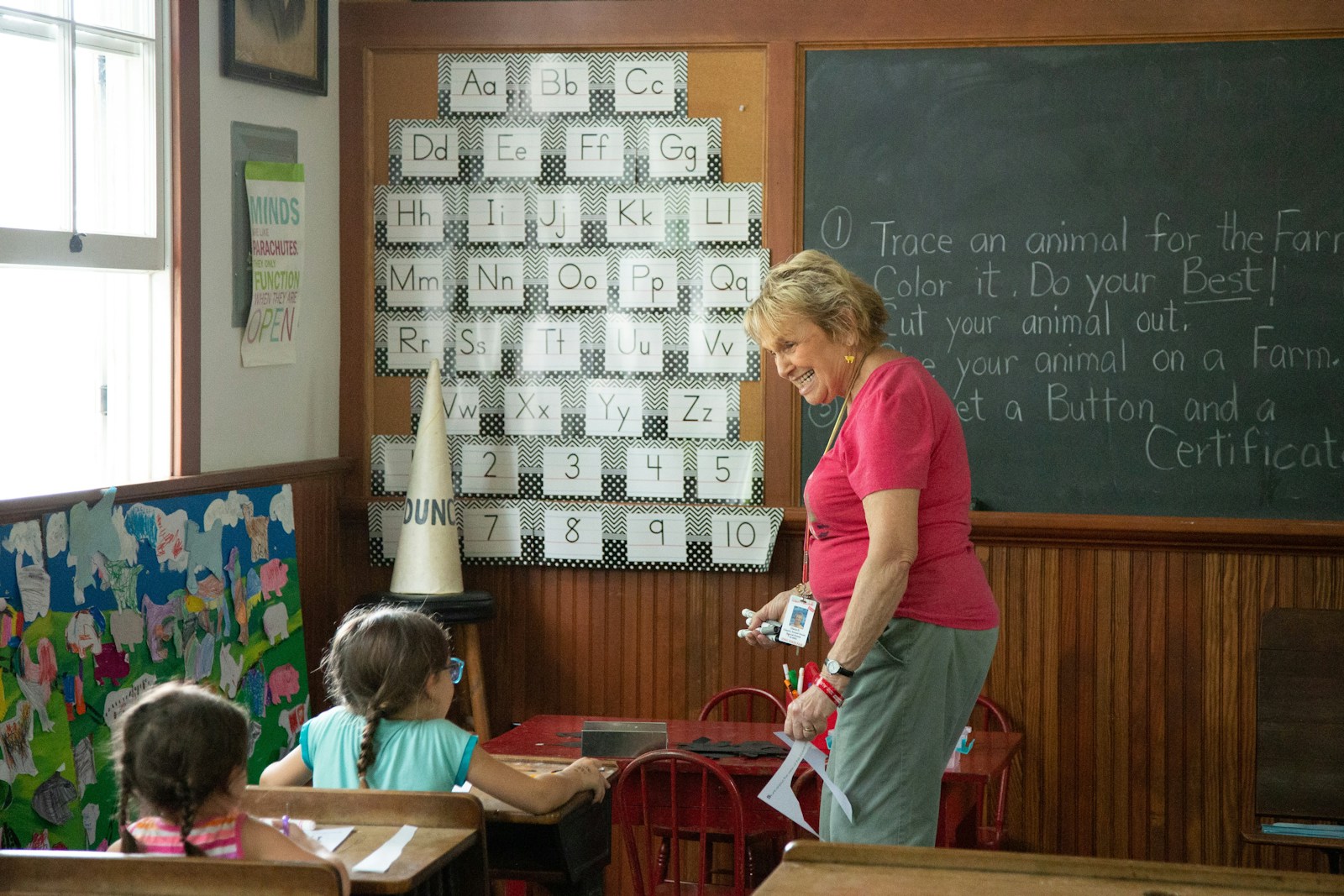 A pre-K teacher in a warm classroom with the full alphabet on the wall behind her, standing with two young students at a small red table.